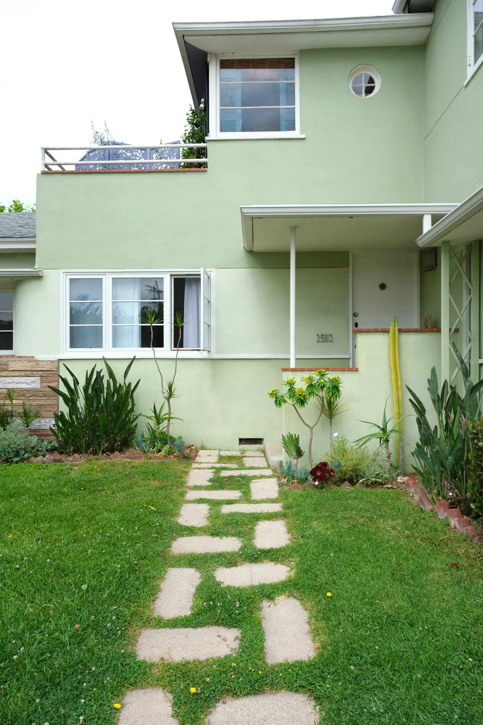 Stepping-stone path across a green front lawn to a mint-green stucco house, succulent border along the foundation
